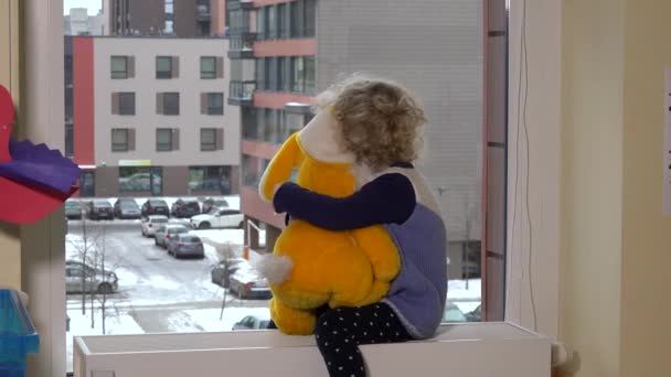 Einsames kleines Mädchen sitzt mit Teddybär am Fenster und betrachtet den Schneefall im Winter — Stockvideo