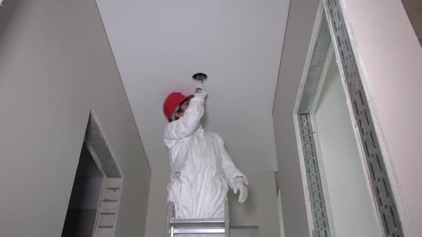 Работник, стоящий на лестнице и прорубающий дыру в потолке коридора для освещения — стоковое видео