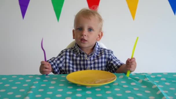 Niño golpeó la mesa con un cuchillo de plástico y tenedor y espera por la comida. Movimiento del cardán — Vídeos de Stock