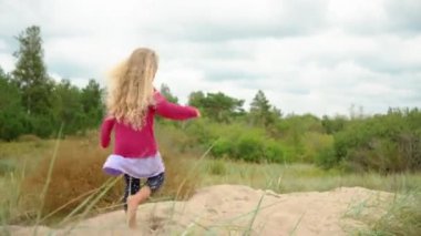 Kum tepesinde çıplak ayakla yürüyen pembe tişörtlü beş yaşında sarışın bir kız.