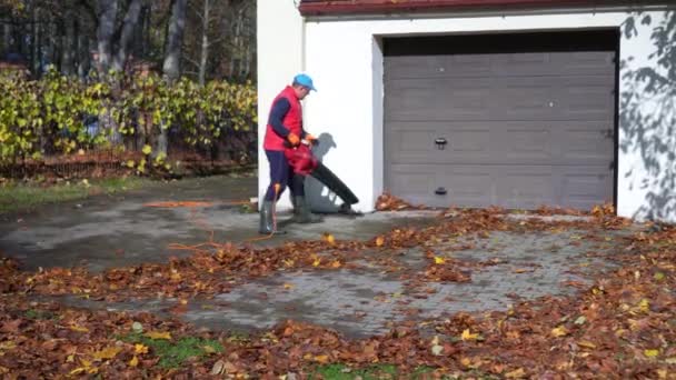 Mannelijke werknemer met bladblazer blaas bladeren in de buurt van garage — Stockvideo