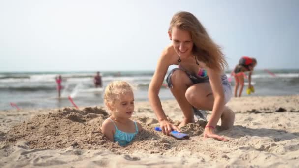 Счастливая женщина и маленькая девочка на пляже. Мама похоронила свою дочь под песком — стоковое видео