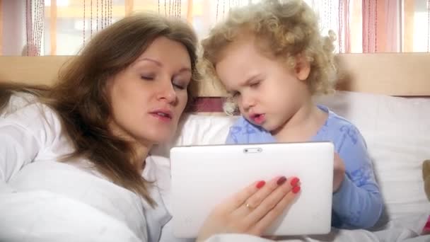 Chica jugar con la tableta y la madre ayudando a jugar. ambos se encuentran en ropa de cama blanca. 4K — Vídeos de Stock