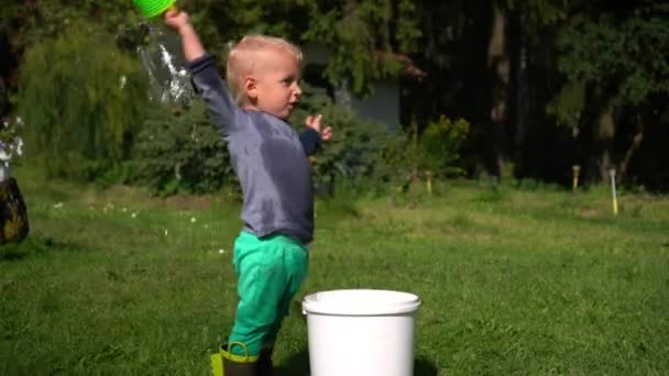 Маленький мокрый мальчик играет с пластиковой лейкой и большим ведром — стоковое видео