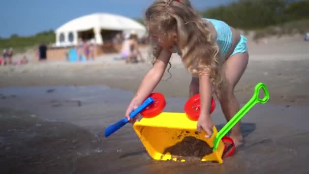 Blondes kleines Mädchen beim Waschen von Spielzeugwagen in den Wellen des Meeres. Liebenswerter Kinderspielstrand — Stockvideo