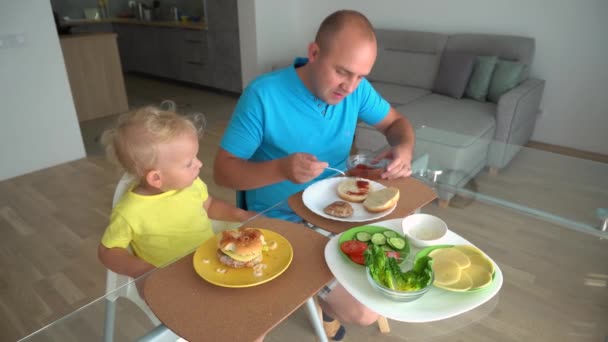 Papai com o filhinho comendo hambúrguer sentado à mesa de vidro. Movimento da câmera Gimbal — Vídeo de Stock