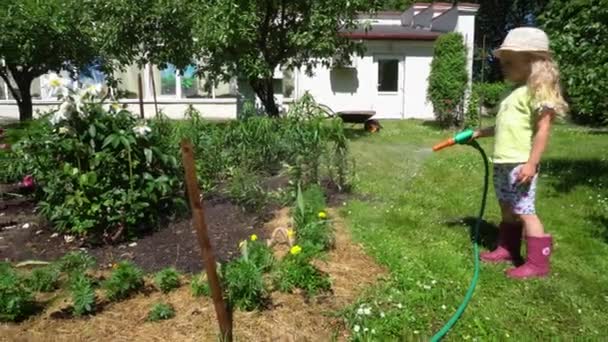 Chica rubia con manguera de agua regando flores en el patio de la casa. Movimiento de cardán — Vídeos de Stock