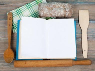 Açık tarif kitabı ahşap arka plan, kaşık, oklava, yeşil kareli masa örtüsü.