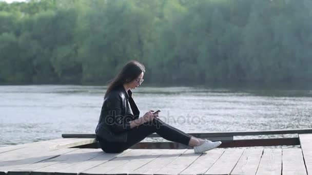 Belle fille est assise sur une rivière et utilise son appareil mobile — Video