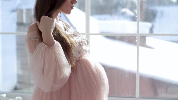 Беременная девушка сидит у окна в красивом розовом платье — стоковое видео