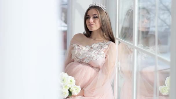 Terhes lány ül egy gyönyörű rózsaszín ruhás ablak közelében — Stock videók