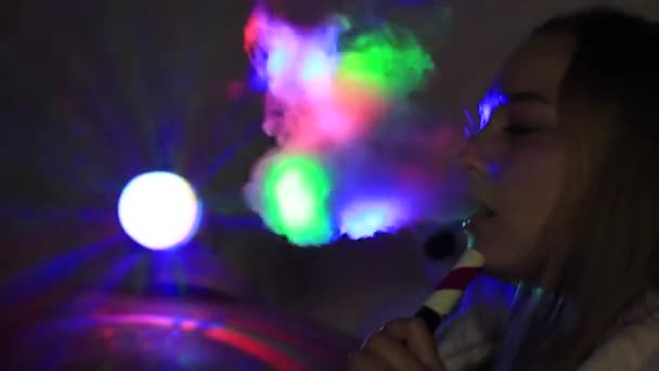 Una chica fuma una cachimba en la oscuridad bajo la luz de una lámpara multicolor . — Vídeos de Stock