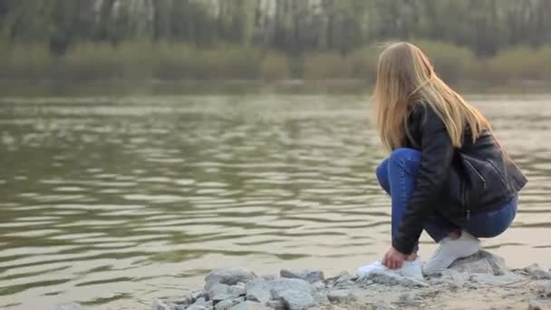 Dziewczyna na brzegu rzeki wrzuca kamień do wody. Widok z tyłu. — Wideo stockowe