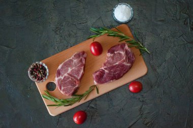 Düz bir zeminde, çiğ et biftekleri, domuz eti, biberiye, deniz tuzu ve domatesler kesme tahtasının üzerinde, koyu gri beton arka planda.