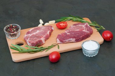 İki çiğ domuz bifteği, deniz tuzu, biberiye, sarımsak, kırmızı domates ve biberler açık ahşap kesim tahtasında, koyu gri beton arka planda.