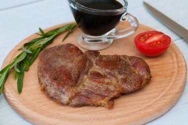 Kızarmış, lezzetli dana eti, biftek ya da domuz eti, domates, biberiye ve soya sosu yuvarlak ahşap bir tahtada, hafif bir arka planda, bıçak ve çatal, yakın plan