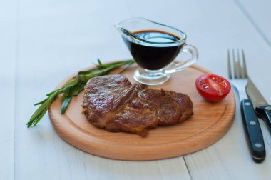Ahşap bir tahtanın üzerinde pişmiş, ızgara domuz eti, sos, biberiye ve domates, çatal ve bıçak, fotokopi alanı olan hafif bir arka planda.