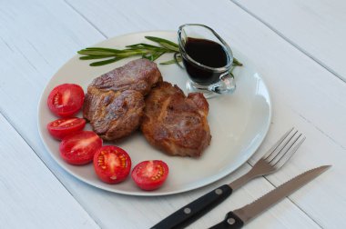 Sos, biberiye ve domatesli gri bir tabakta leziz ızgara domuz bifteği. Hafif ahşap arka planda, çatal bıçak takımında.