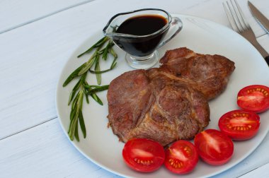 Sos, biberiye ve domatesli gri bir tabakta leziz ızgara domuz bifteği. Hafif ahşap arka planda, çatal bıçak takımında.