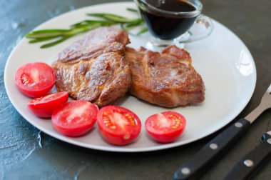 Baharatlı, domatesli ve soslu kokulu domuz eti bifteği gri bir tabakta, metin için yeri olan koyu beton arka planda.
