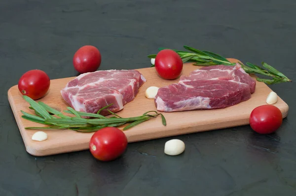 Filetes de cerdo crudos con tomates rojos, romero y ajo en una tabla de cortar . — Foto de Stock