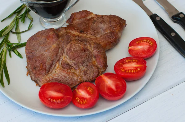 Ízletes, grillezett sertéshús szürke tálcán, mártással, rozmaringgal és paradicsommal, világos fa alapon, evőeszközökkel, közelről — Stock Fotó