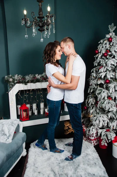 Belo casal apaixonado, homem e mulher, abraçando e olhando um para o outro no interior do Ano Novo perto da árvore de Natal e lareira — Fotografia de Stock