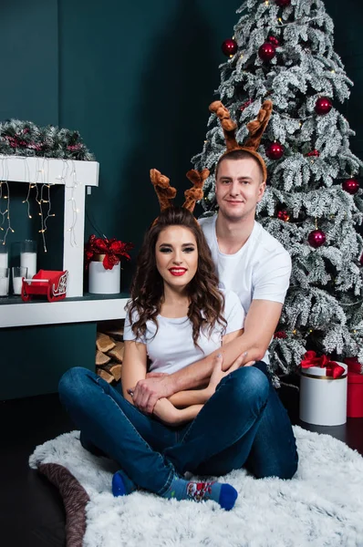 一对快乐的年轻夫妇，头上顶着鹿角，靠近壁炉和圣诞树 — 图库照片