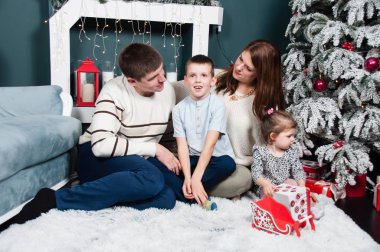Aile, aileler ve iki çocuk, kız ve erkek, gülümsüyor, halıdaki bir Noel ağacının şöminesinin yanında eğleniyorlar.