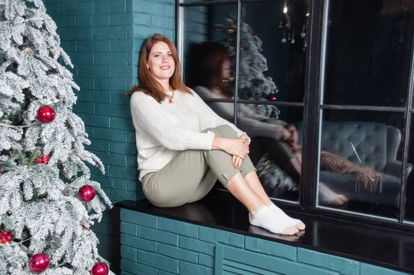 Uma mulher muito bonita, feliz e misteriosamente sorridente senta-se no parapeito da janela perto de uma árvore de Natal . — Fotografia de Stock