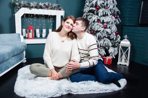 可爱的夫妻,英俊的男人和美丽的女人在圣诞树边和壁炉边. 丈夫亲吻他妻子的脸颊 — 图库照片