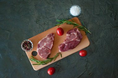 Düz bir zeminde, çiğ et biftekleri, domuz eti, biberiye, deniz tuzu ve domatesler kesme tahtasının üzerinde, koyu gri beton arka planda.