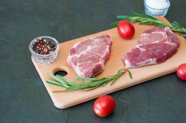 Çiğ biftek, domuz eti, biber, biberiye, deniz tuzu ve domatesler kesme tahtasının üzerinde, koyu gri beton arka planda, yakın plan