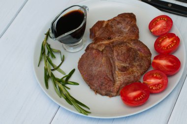 Sos, biberiye ve domatesli gri bir tabakta leziz, ızgara domuz bifteği, hafif ahşap arka plan, çatal bıçak takımı, yakın çekim.