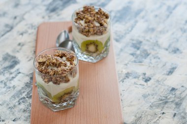 Granola, kivi, muz ve Yunan yoğurt ahşap tahta gri beton üzerinde iki cam bardak, kaşık, kopya alanı ile, üstten ve yandan görünümü. Fitness, şekil, vücut ve sağlıklı gıda
