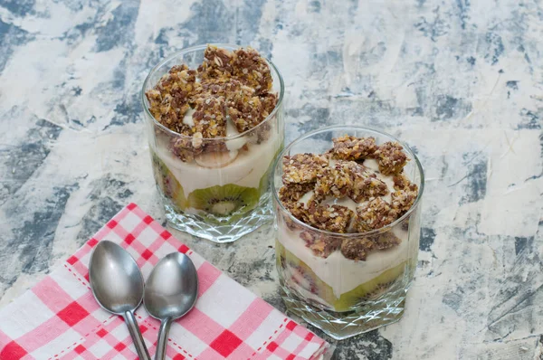 Dessert granola, Greek yogurt, kiwi and banana in two glass cups with spoons and pink checkered towel on gray concrete. Fitness, figure, body and healthy food — Stock Photo, Image