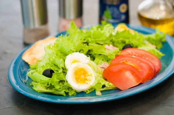 Salát s tuňákem, vejci, rajčaty, olivami, kořením a krutony, omáčkou nebo olejem na modrém talíři — Stock fotografie
