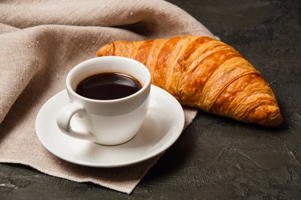 Croissant et tasse de café et soucoupe sur fond sombre avec une serviette en lin gris — Photo
