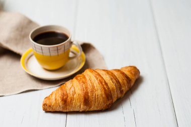 Klasik sarı bardakta espresso, çay tabağı ve kaşık, keten peçeteli kruvasan, hafif arka plan, öğle yemeği konsepti.