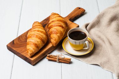 Kaliteli sarı fincanda kahve, çay tabağı ve kruvasan, ahşap tabakta keten peçete, arka planda hafif bir kahvaltı konsepti.