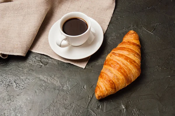 Круассан и чашка кофе и блюдце на темном фоне с серой салфеткой — стоковое фото