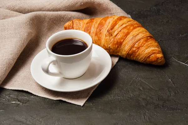 Круассан и чашка кофе и блюдце на темном фоне с серой салфеткой — стоковое фото