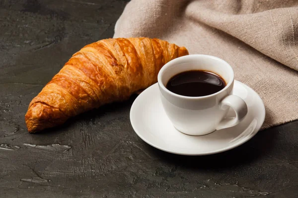 Croissant et tasse de café et soucoupe sur fond sombre avec une serviette en lin gris — Photo
