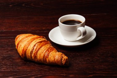 Kruvasan ve kahve, koyu ahşap masa üstünde beyaz bardakta. Masada kahvaltı konsepti
