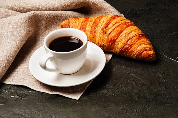 Croissant sucré savoureux, tasse blanche de café ristretto fort sur une soucoupe et une serviette de lin sur un fond sombre — Photo