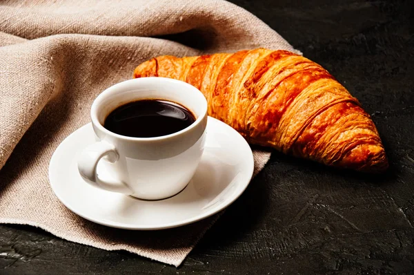 Croissant sucré savoureux, tasse blanche de café ristretto fort sur une soucoupe et une serviette de lin sur un fond sombre — Photo