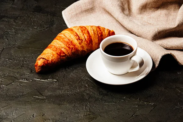 Croissant sucré savoureux, tasse blanche de café ristretto fort sur une soucoupe et une serviette de lin sur un fond sombre avec espace de copie — Photo