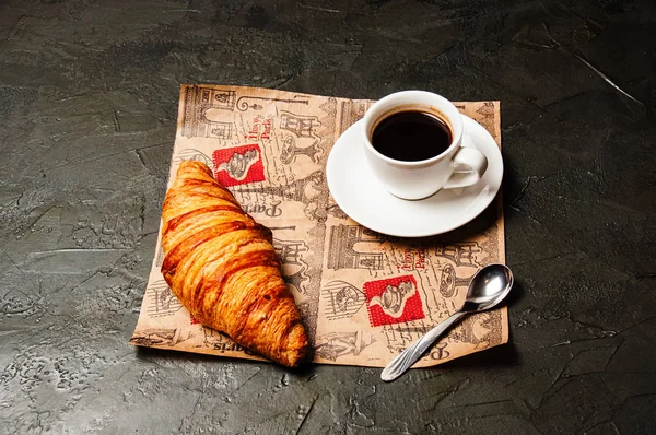Французский круассан, сладкий крепкий кофе в белой чашке на блюдце с ложкой, на красивой бумаге ремесла, на темном фоне — стоковое фото