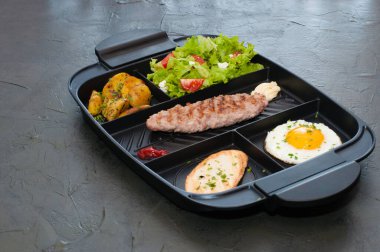 Kahvaltı, pirzola ya da biftek, domates salatası, kızarmış patates, kızarmış ekmek ve siyah tabakta kızarmış yumurta. Restoran yemeği, site için içerik