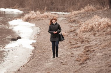 Kıvırcık kızıl saçlı çok güzel bir kız bulutlu bahar havasında nehir kıyısında yürüyor.